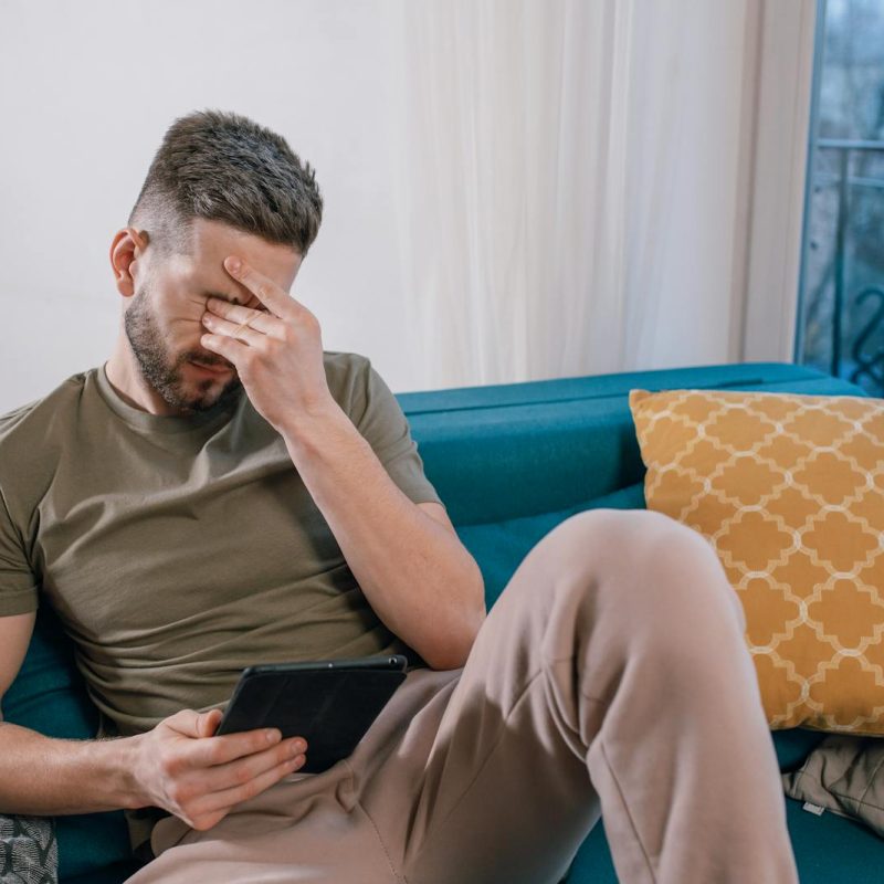 A man sits on a sofa holding a tablet, visibly stressed and covering his face with one hand
