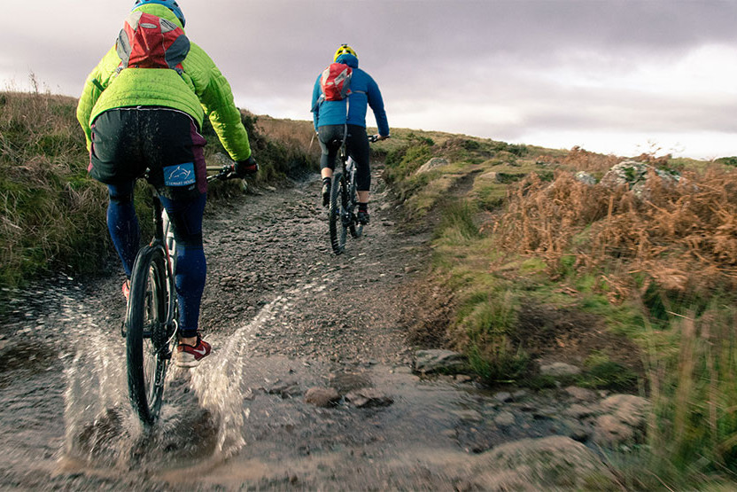 Two individuals cycling on a muddy trail, surrounded by greenery, showcasing an adventurous outdoor activity